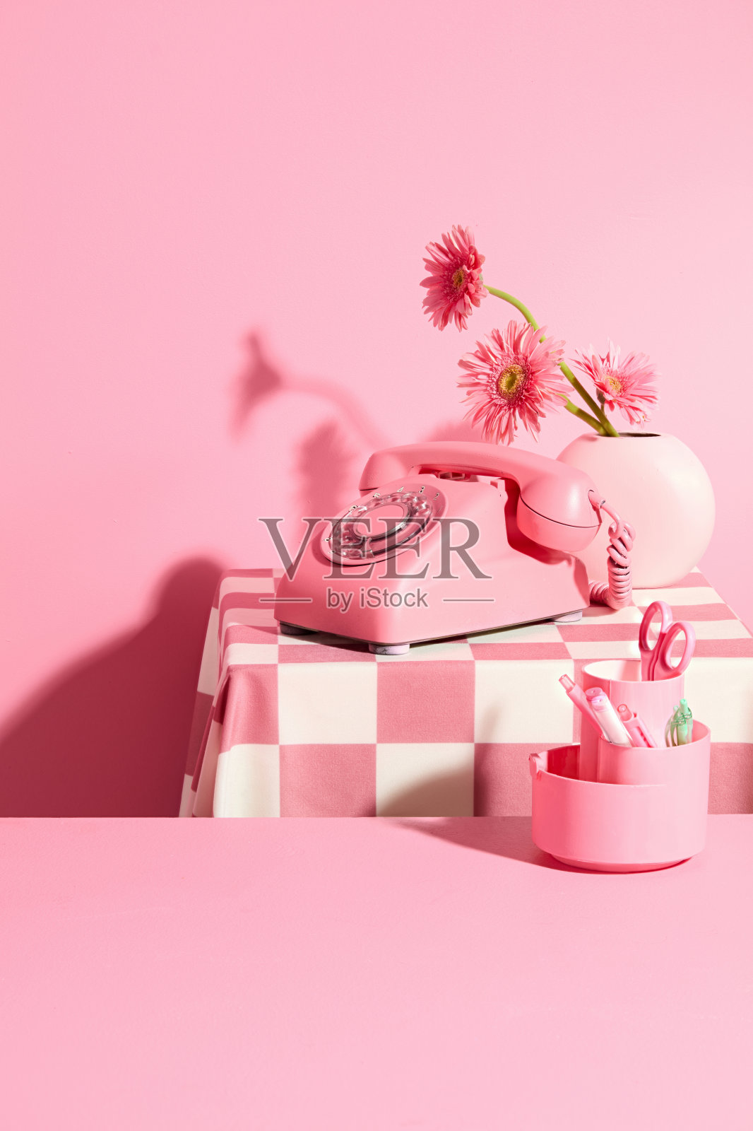 一张粉红色的空桌子上装饰着一盒办公用品，后面是一张铺着粉红色桌布的桌子，桌子上放着一台台式电话和一瓶非洲菊。中心有空位供展示照片摄影图片