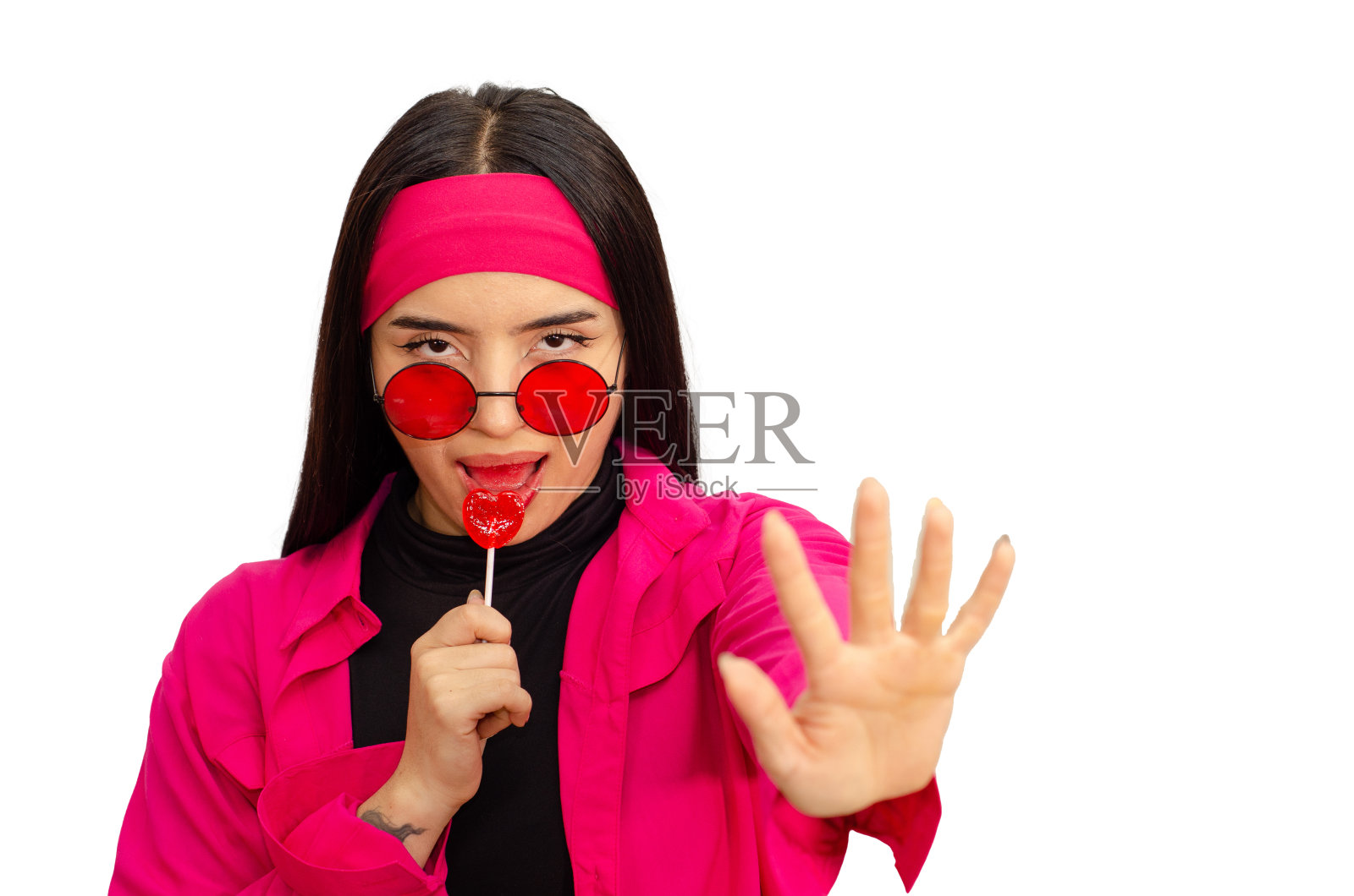 美丽而快乐的年轻女子的肖像，穿着休闲的衣服，戴着发带和红色太阳镜，吃着白色背景的心形棒棒糖照片摄影图片