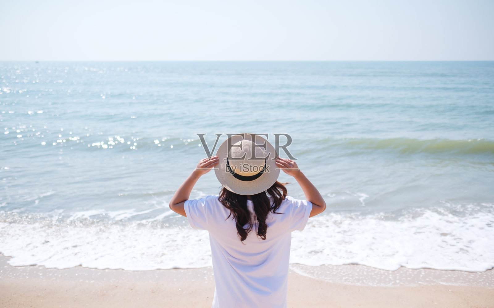 一个戴着帽子的女人站在蓝天背景的沙滩上的后视图照片摄影图片