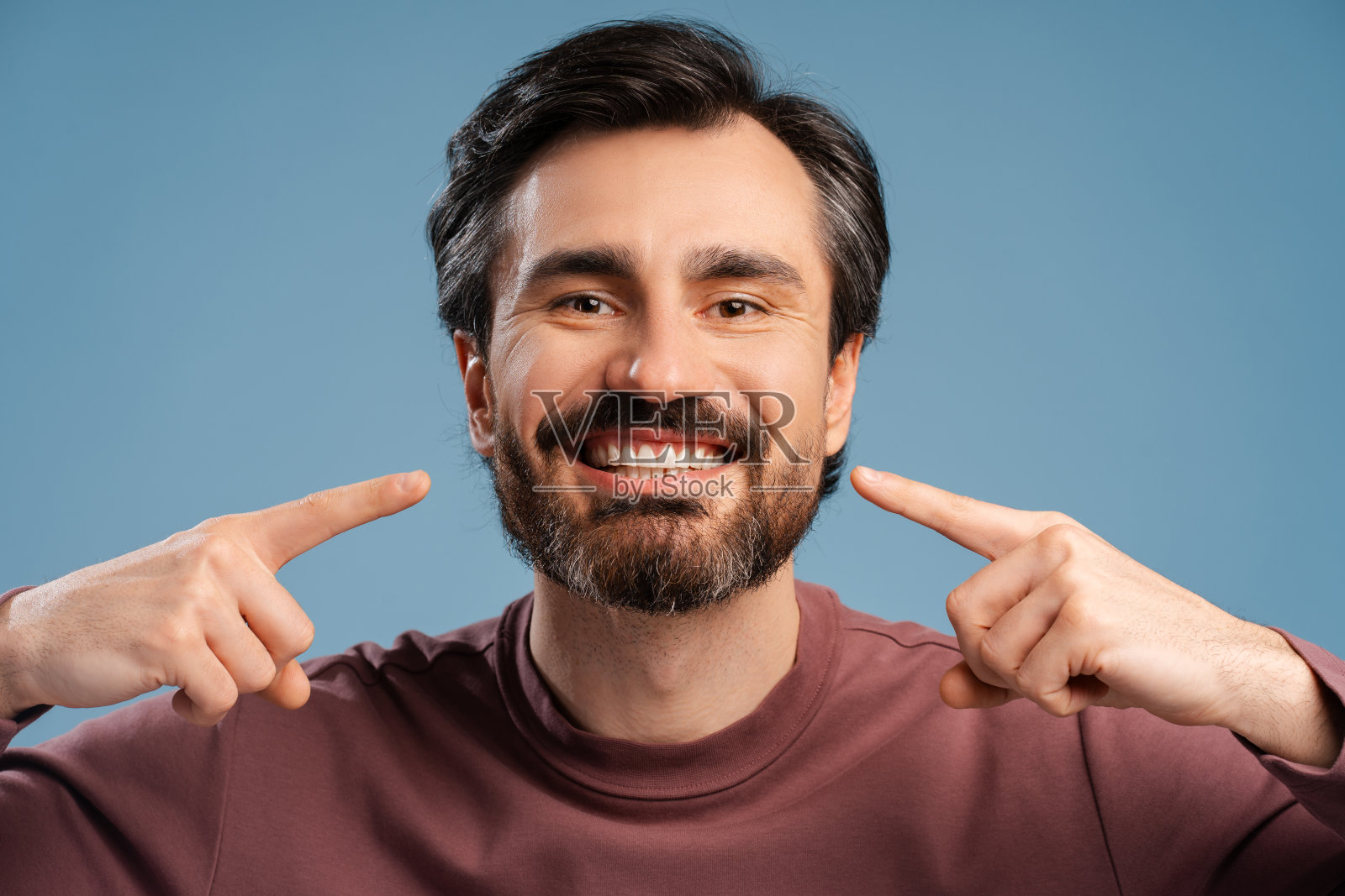 快乐的成年大胡子男人指着牙齿微笑孤立在蓝色工作室照片摄影图片