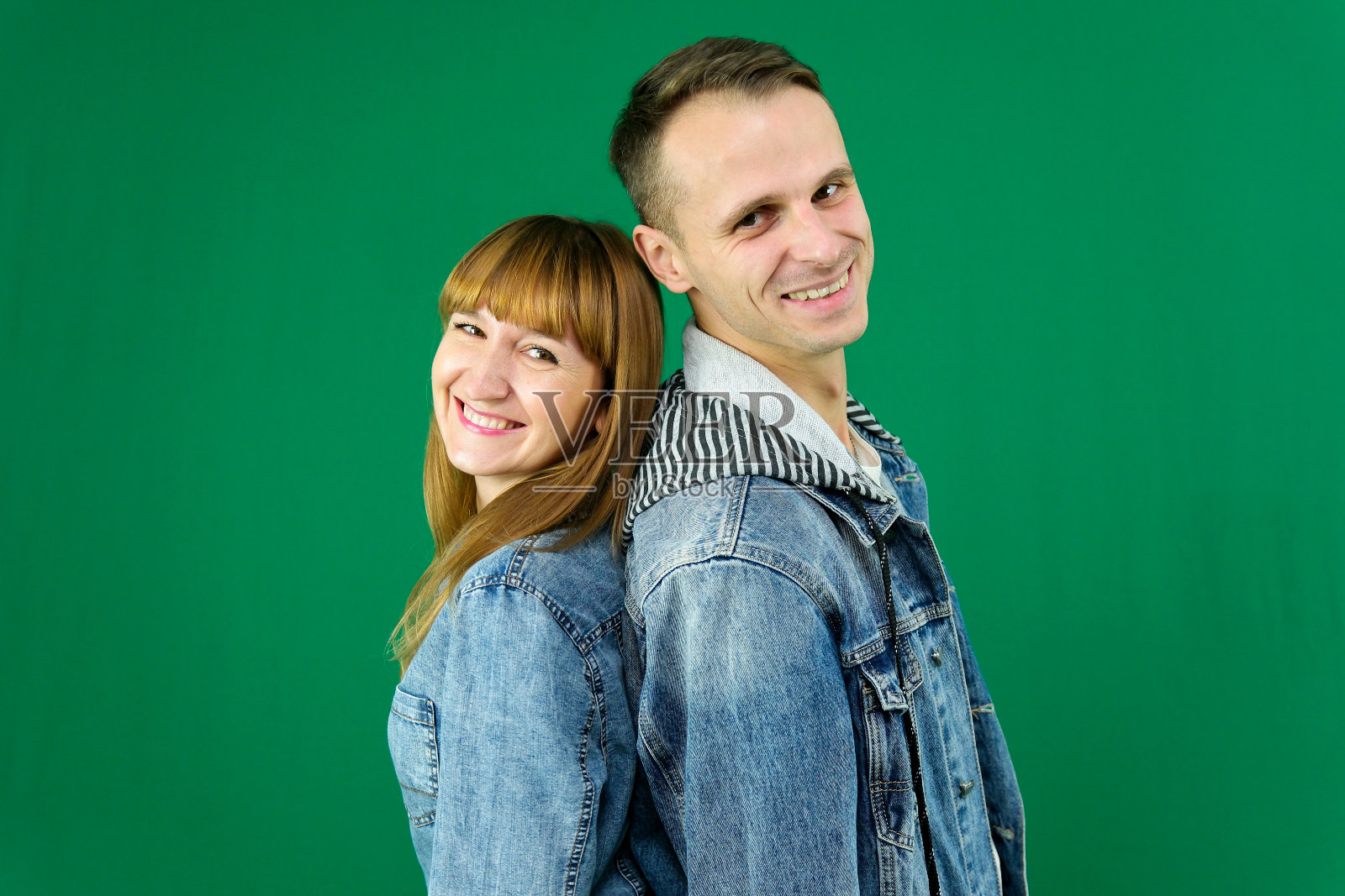 年轻欢快的夫妇微笑着，拥抱着，穿着，背对着对方站着，绿色背景上的牛仔衣服照片摄影图片