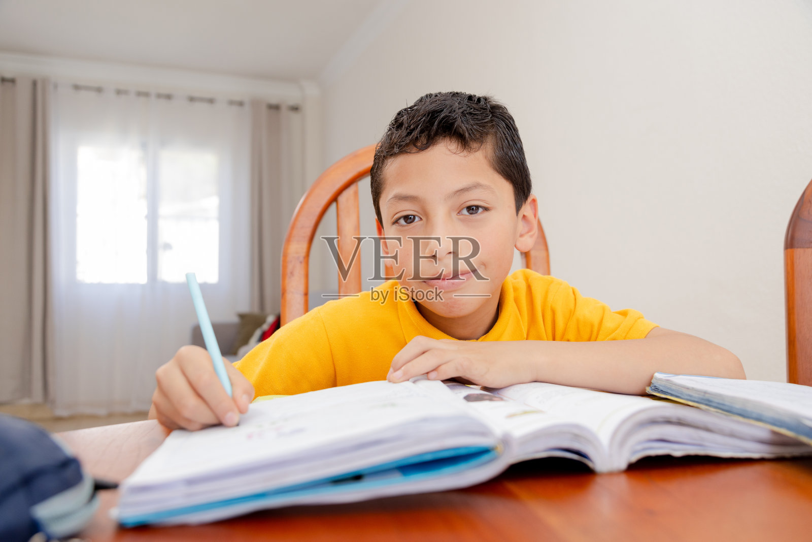 西班牙男孩在家做作业-穿黄色t恤的男孩在笔记本上写字-家庭学校- 8岁的男孩在学习。西班牙男孩一边对着镜头微笑一边做作业——穿黄色t恤的男孩在他的书上写字——家庭学校——8照片摄影图片