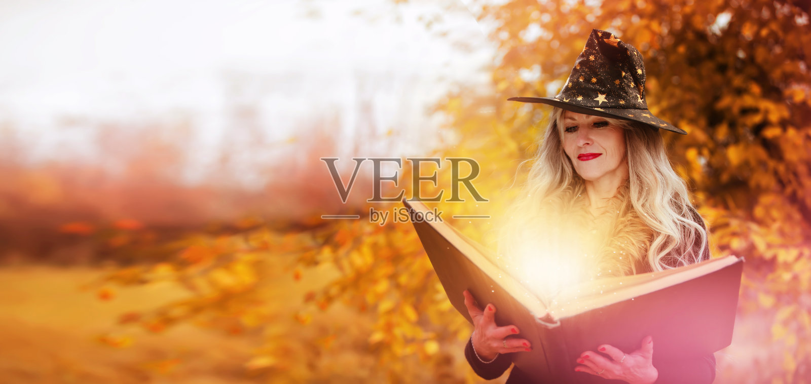 万圣节前夕，一个戴着女巫帽、拿着魔法书的女人正在变魔术，在秋天的森林背景上表演巫术。照片摄影图片