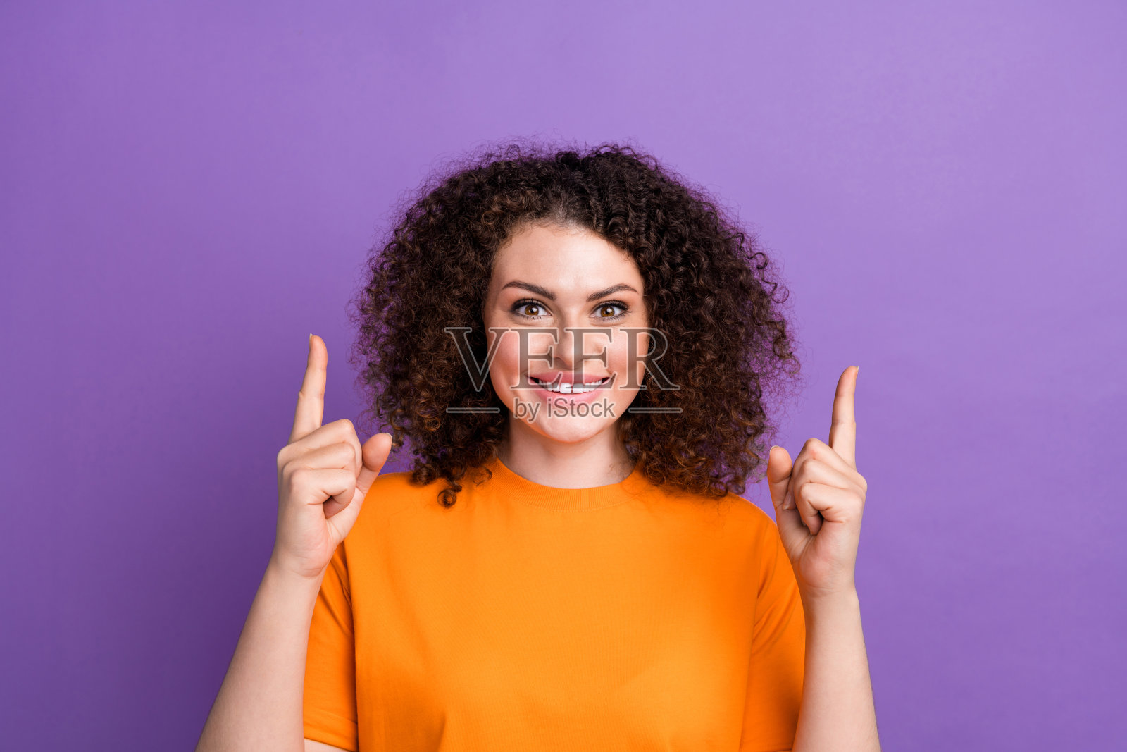 照片中穿着橙色t恤的时髦女士兴奋地用两个手指指着空的空间，孤立的紫色背景照片摄影图片