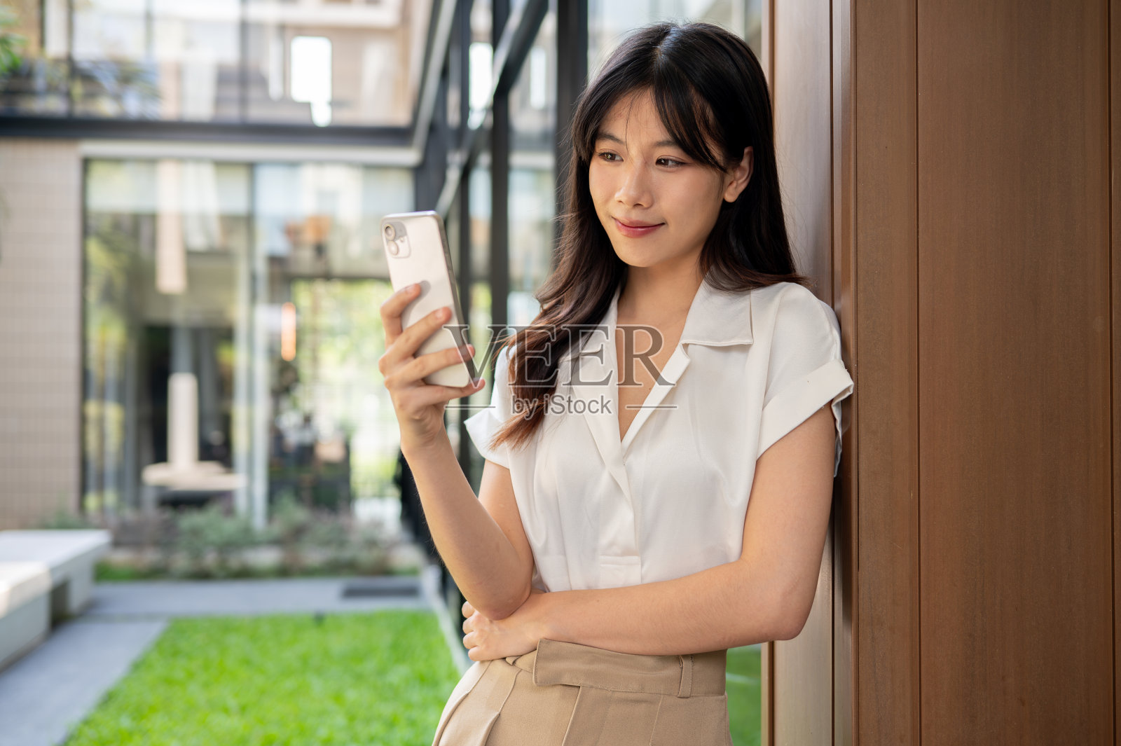 一位迷人的亚洲女商人倚在大楼外的一堵墙上使用她的智能手机。照片摄影图片