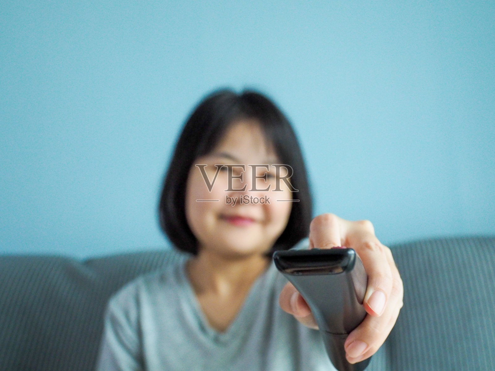年轻女子拿着遥控器，微笑着，兴奋地坐在家里的沙发上看电视。照片摄影图片