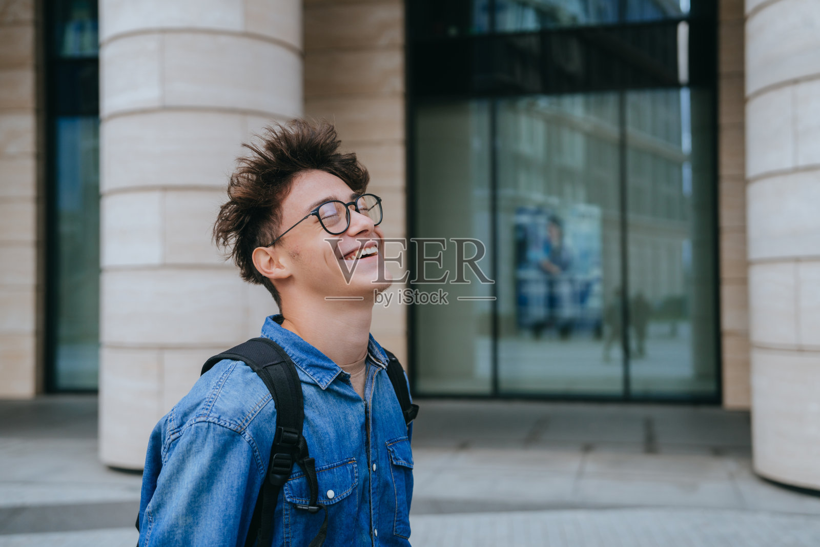 背着背包走在街上笑着的少年去上大学了。戴眼镜的帅气男生笑得睁大眼睛。教育，大学生活。照片摄影图片