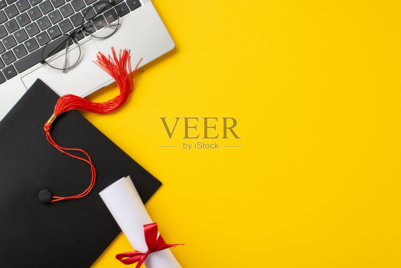 这是一个充满活力的场景，带有红色流苏的毕业帽、文凭、眼镜和黄色背景上的笔记本电脑，象征着在线教育和毕业成就照片摄影图片