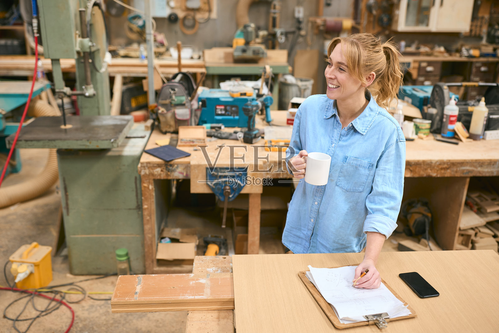 在木工车间工作的女木匠拿着一杯咖啡在剪贴板上做笔记照片摄影图片
