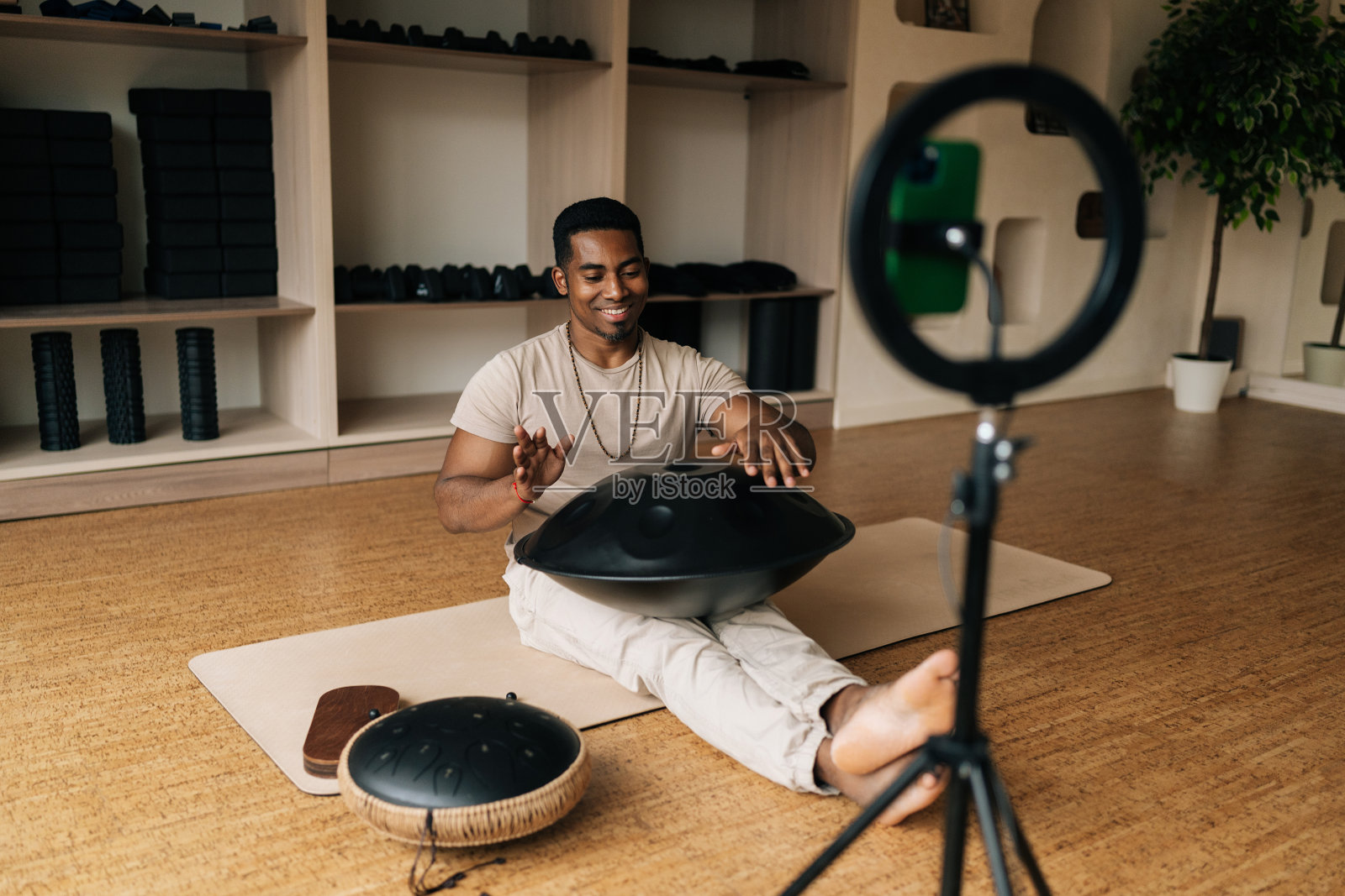 快乐的黑人男博主坐在地板上练习本土音乐传统的鼓乐器，手机上有戒指灯。照片摄影图片