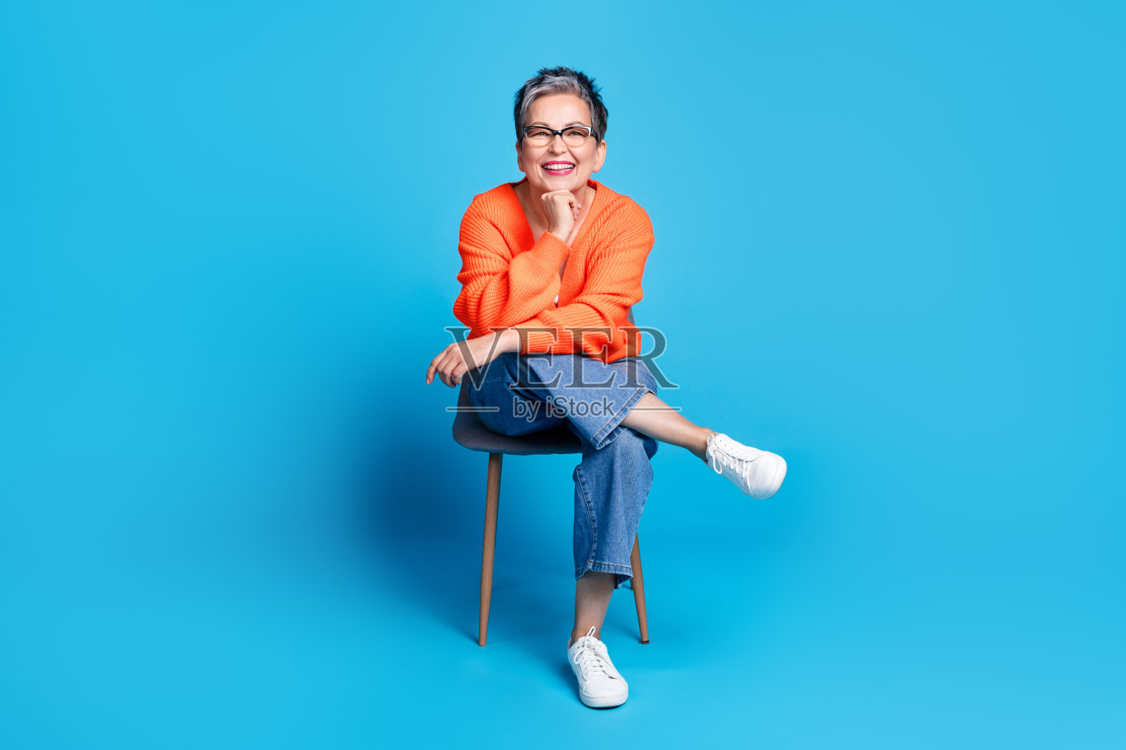 可爱的资深女士的全身照片，坐在椅子上，穿着时尚的橙色服装，孤立在蓝色背景上照片摄影图片