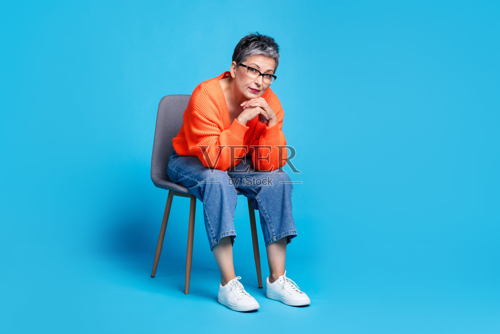 可爱的高级女士的全身照片，坐在椅子上周到的穿着时尚的橙色服装孤立的蓝色背景照片摄影图片