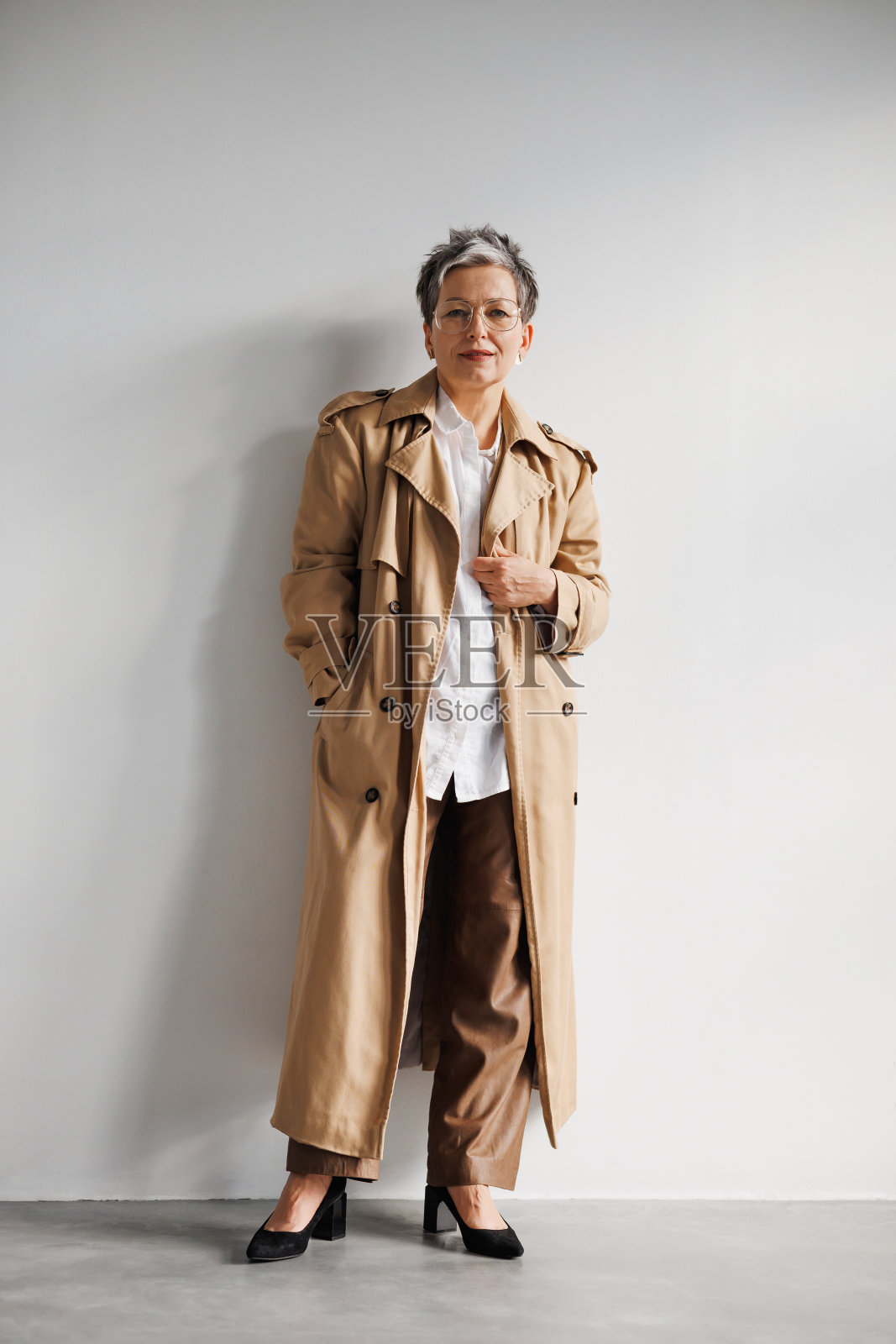 自信优雅的成熟女人，穿着风衣，戴着眼镜，站在灰色的墙边照片摄影图片