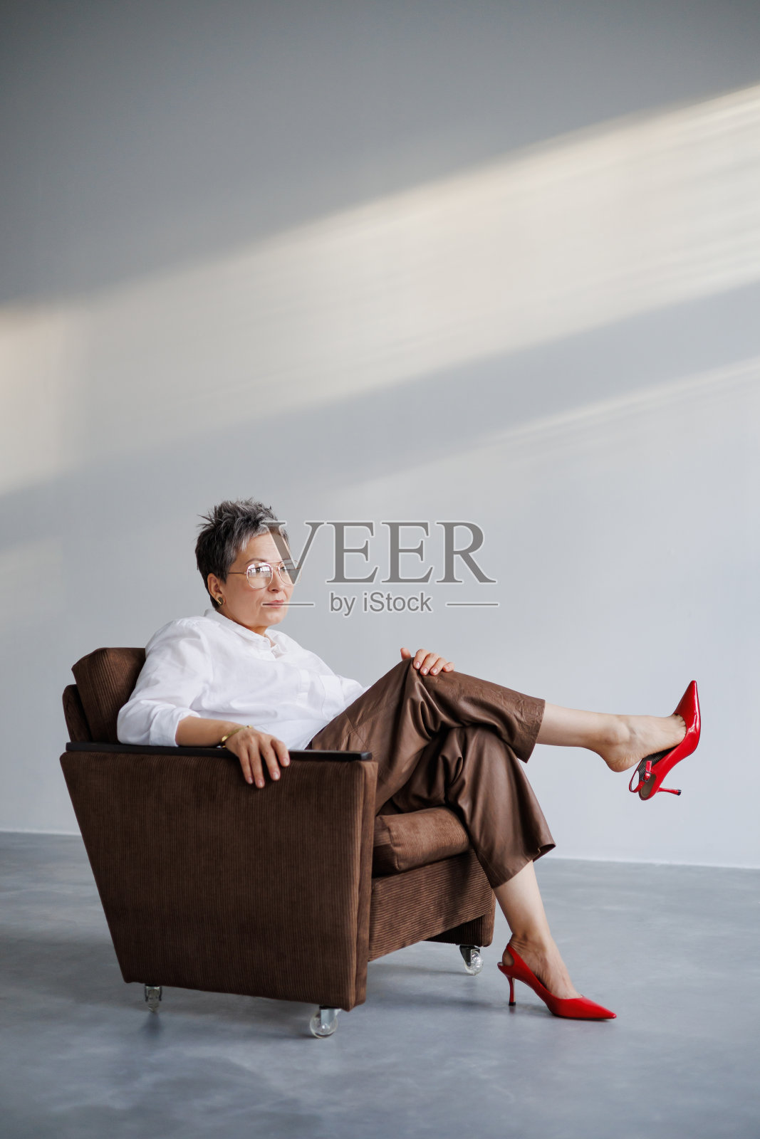 时尚成熟的女人穿着红高跟鞋和白衬衫，坐在灰色背景的扶手椅上照片摄影图片