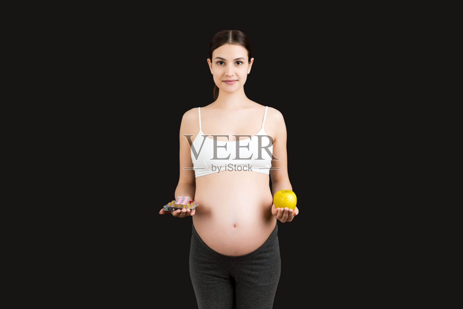 一位孕妇在五彩缤纷的背景下，在苹果和一堆药片之间做出选择。怀孕期间服用维生素照片摄影图片