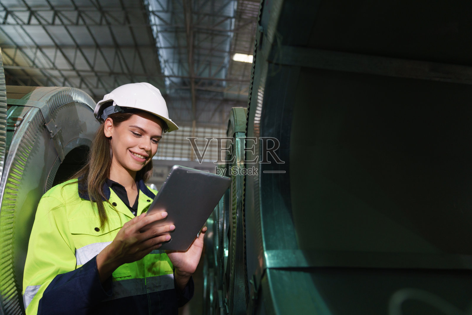 身穿绿色制服、头戴安全头盔的女工在一家金属板材厂内进行质量检查。照片摄影图片