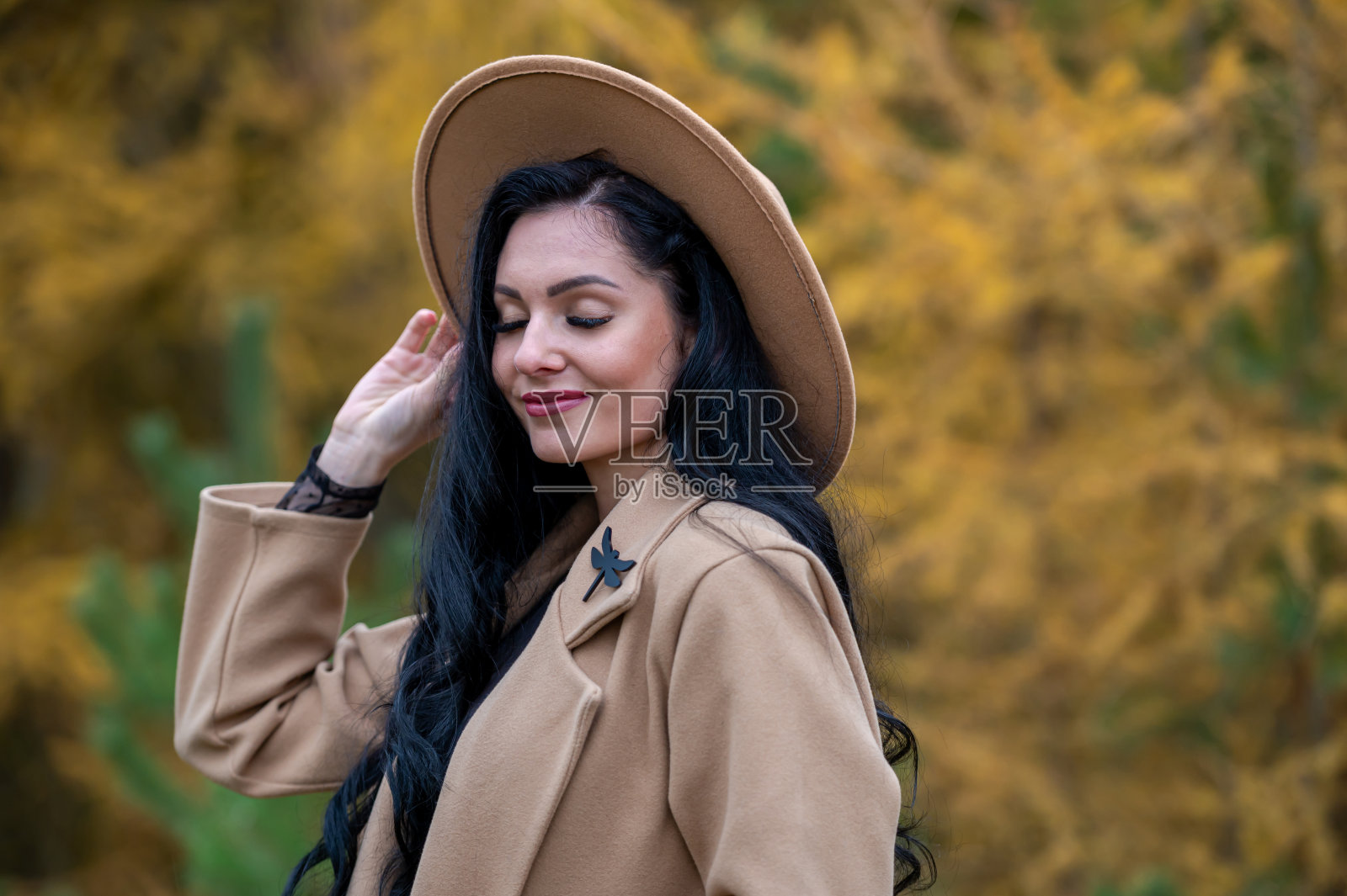 美丽的黑发女孩穿着米色外套，戴着帽子，在秋天的公园里摆姿势照片摄影图片