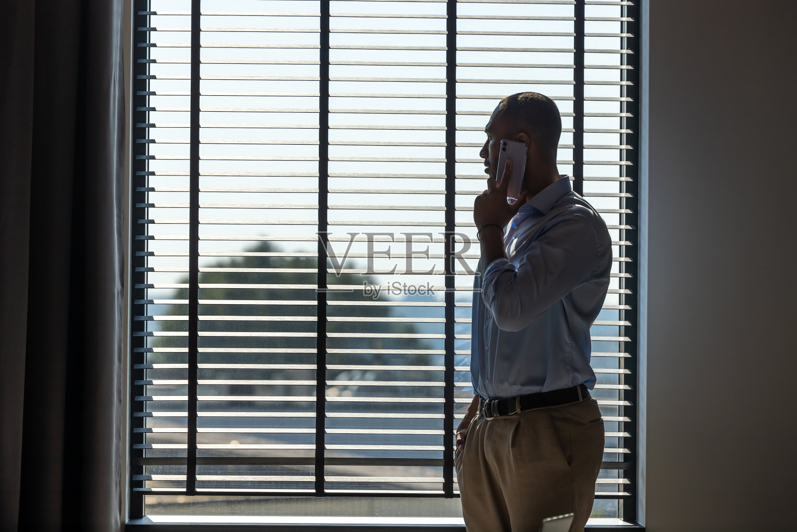 商人站在办公室的大窗户旁边，自信地讲着手机照片摄影图片