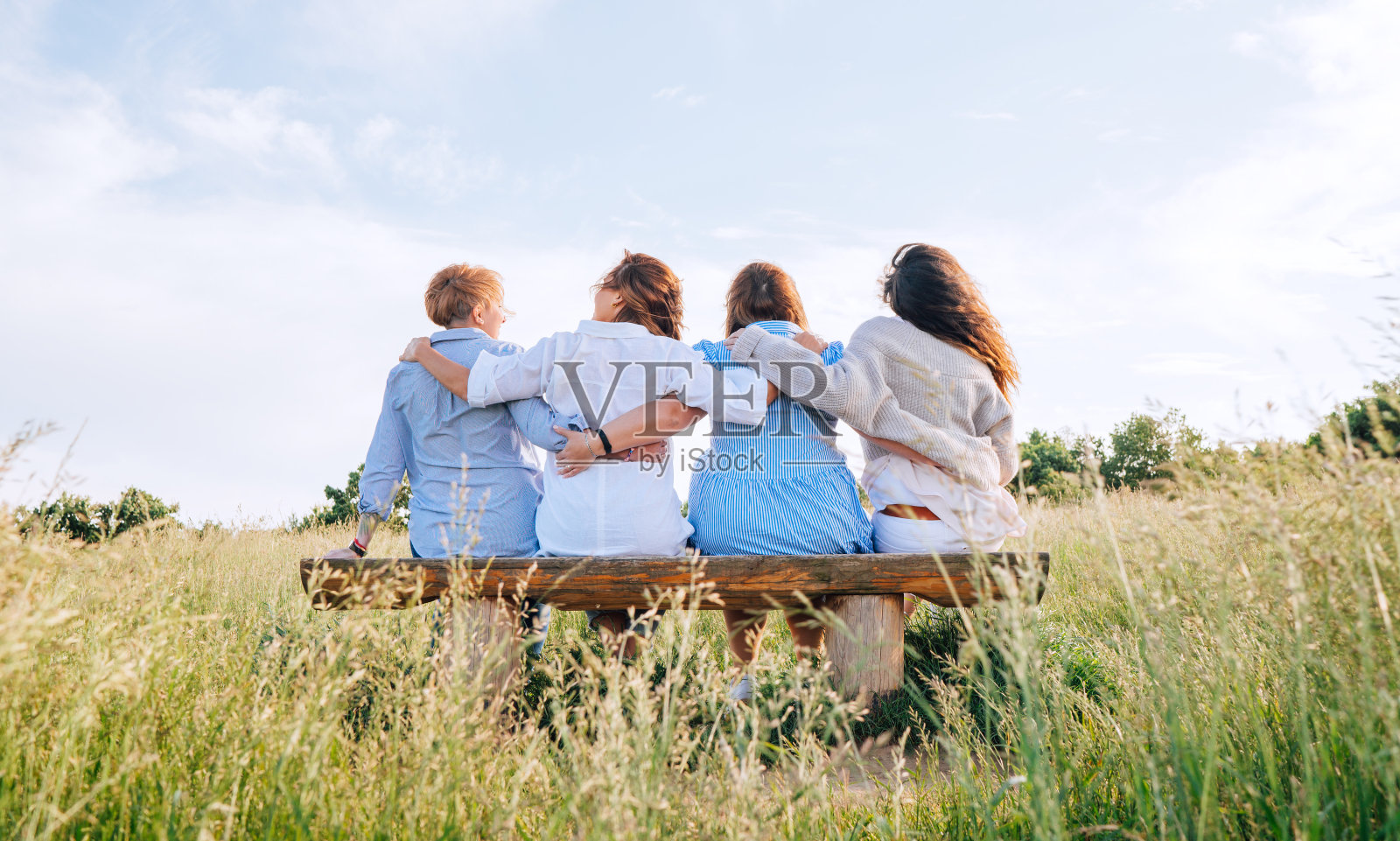 四个面带微笑的妇女穿着轻薄的夏装，在户外散步时坐在草地长凳上，互相拥抱。女人的友谊、关系和幸福概念形象。照片摄影图片