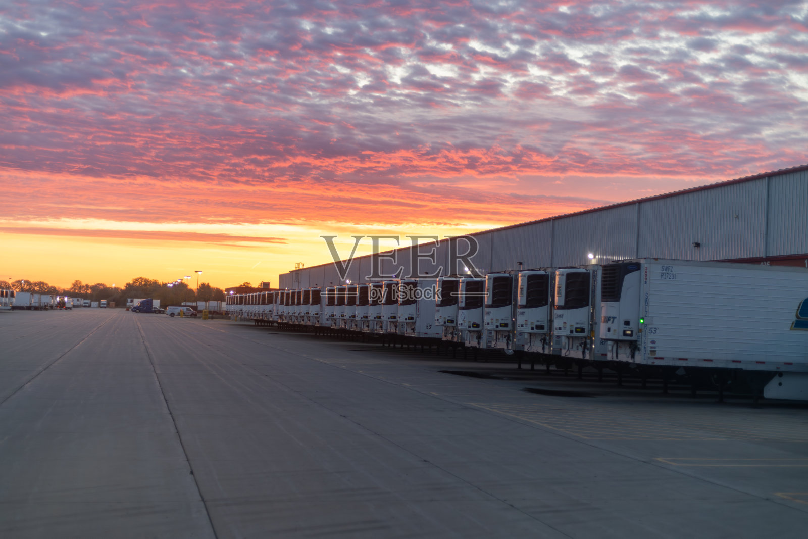 在一个物流园区，一长排白色半挂车停在耀眼的粉色和橙色的日出下。照片摄影图片