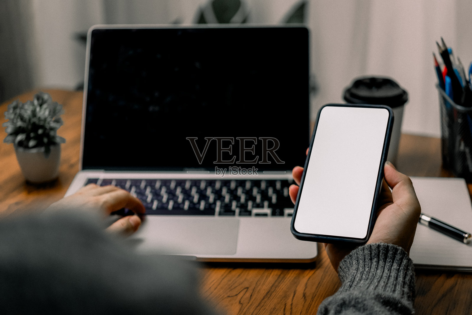 坐在办公桌前使用智能手机和笔记本电脑的人，暗示多任务处理或移动通信。智能手机屏幕一片空白。照片摄影图片