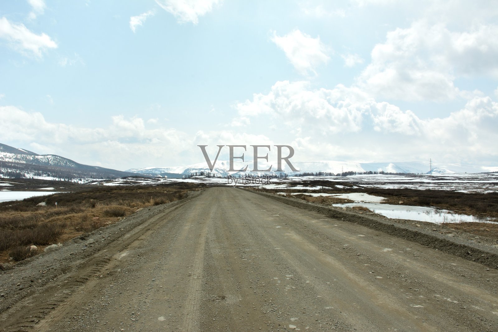 在多云的春日天空下，一条泥泞的砾石路穿过一大片白雪覆盖的空地，四周环绕着高山。照片摄影图片