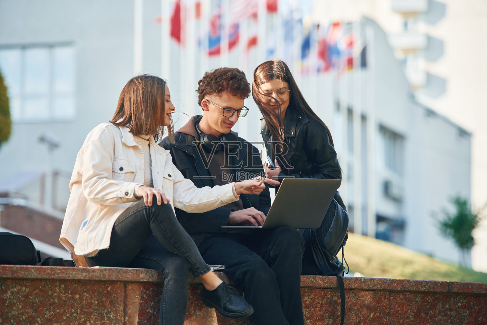 坐着用笔记本电脑。三个年轻的学生在大学外面照片摄影图片