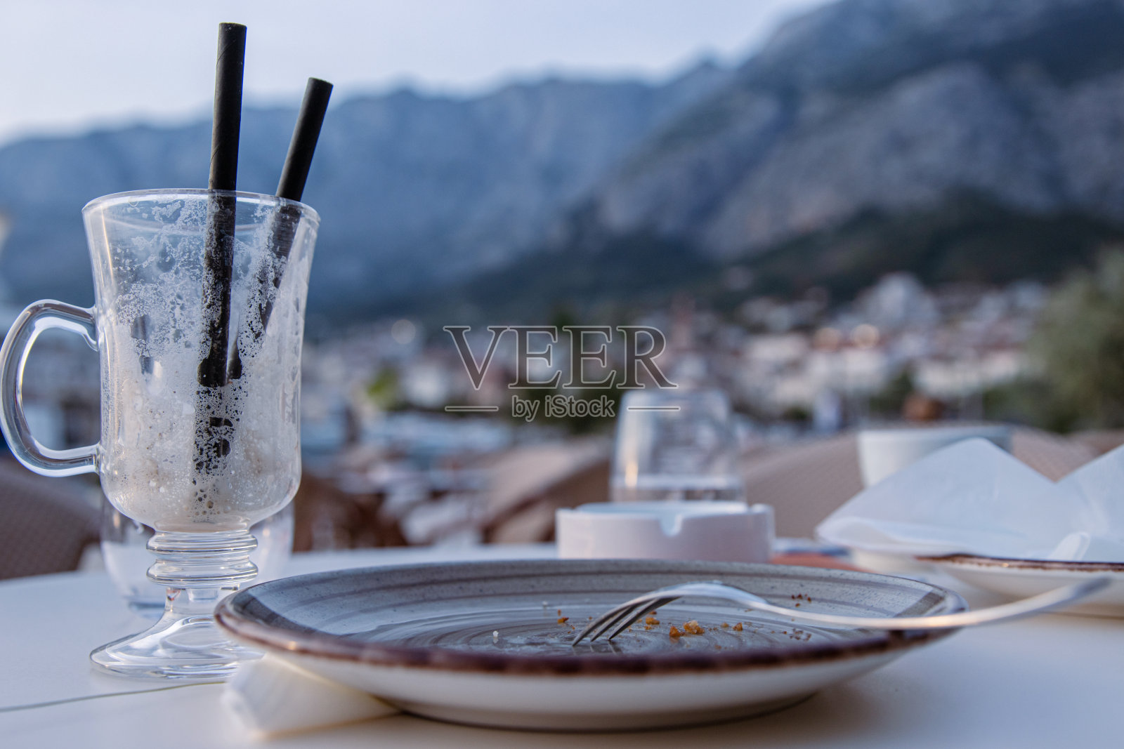 街边咖啡馆桌子上的成品咖啡拿铁或爱尔兰咖啡杯，两根吸管，带叉子的空盘子，烟灰缸，脏盘子和餐巾。侧视图。有选择性的重点。照片摄影图片