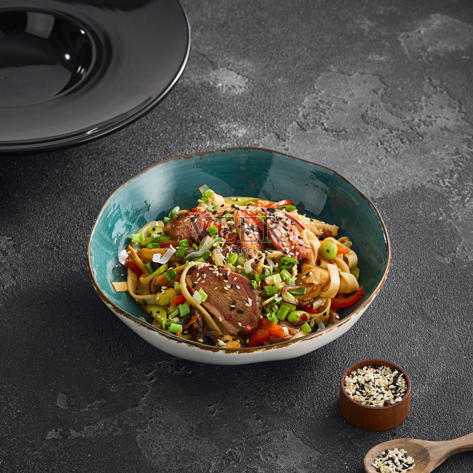 美味的乌冬面与多汁的鸭片和五颜六色的蔬菜一起炒，放在一个黑色纹理表面的碗里照片摄影图片