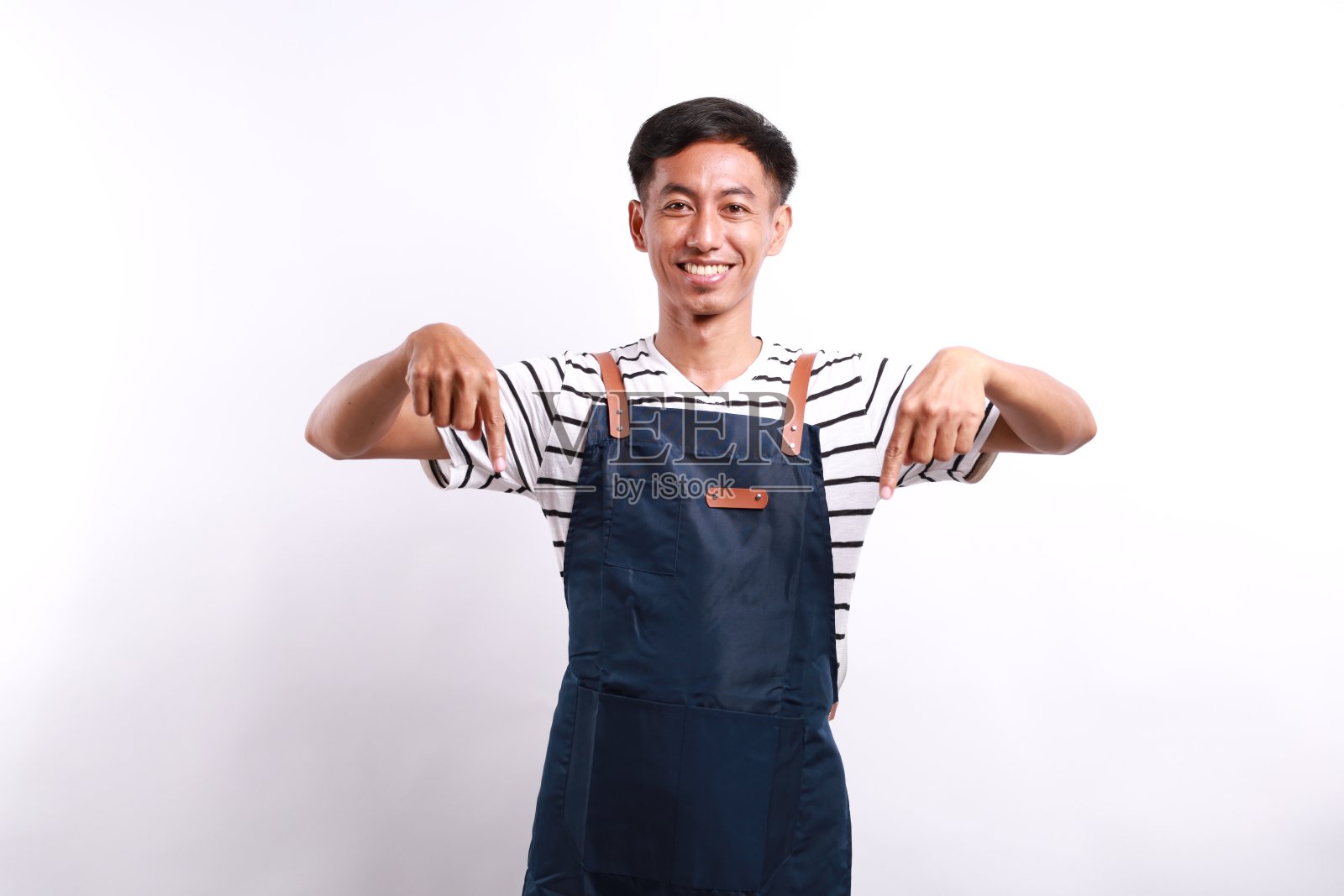 穿着蓝围裙的友好的亚洲咖啡师指着下面，展示你的标志横幅，站在白色的背景上照片摄影图片