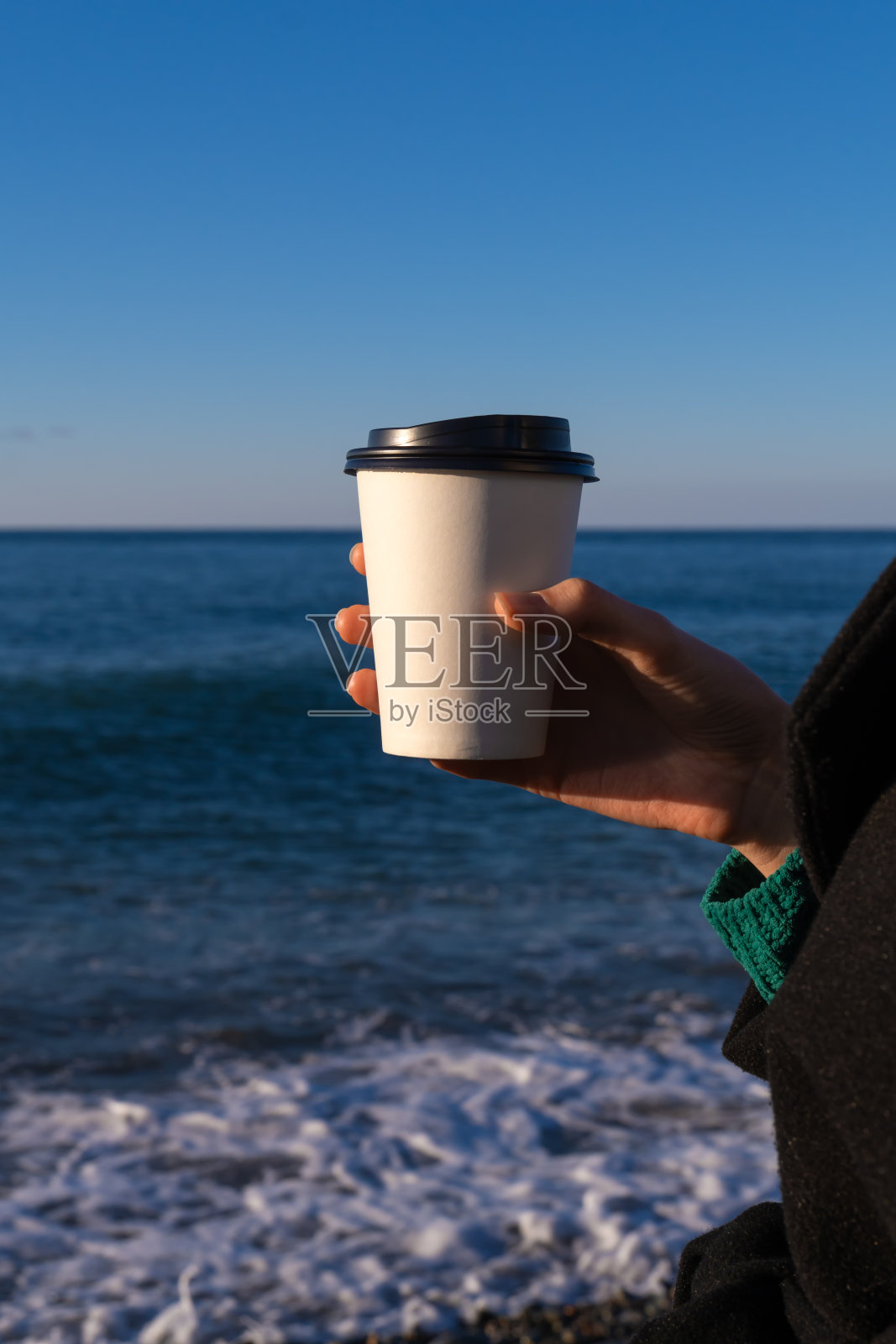在一个阳光明媚的日子里，一个女人的手拿着一个装有咖啡或茶的白色一次性纸杯，以大海为背景的特写，复制空间。垂直图照片摄影图片