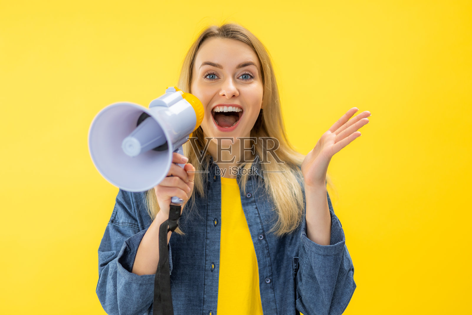 微笑的年轻女子对着扩音器尖叫，独自在黄色背景的摄影棚里摆姿势照片摄影图片