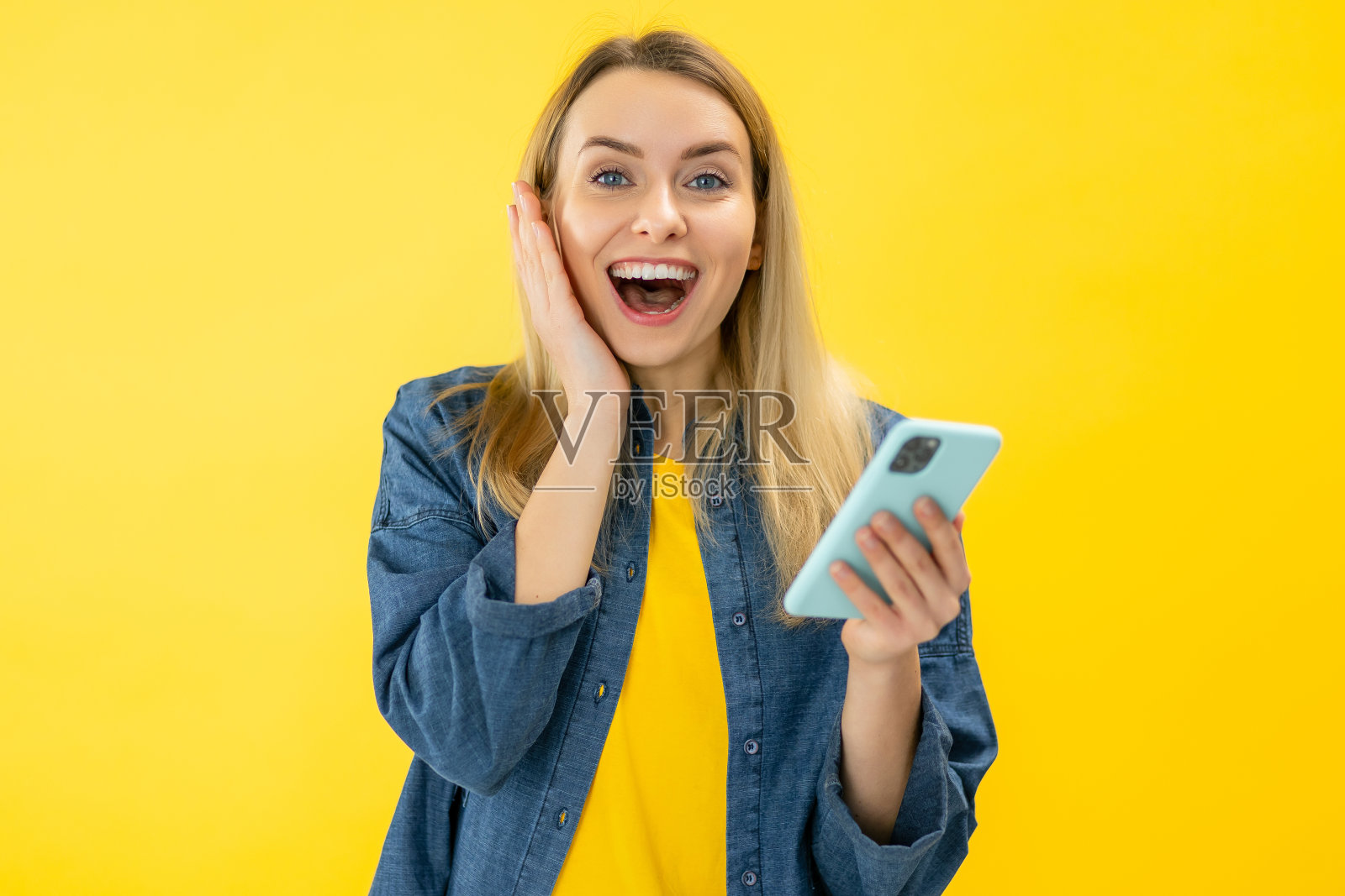 快乐满足的女人拿着智能手机，微笑着发出哇，庆祝在线彩票或赠送胜利，在黄色背景下独自下注。照片摄影图片