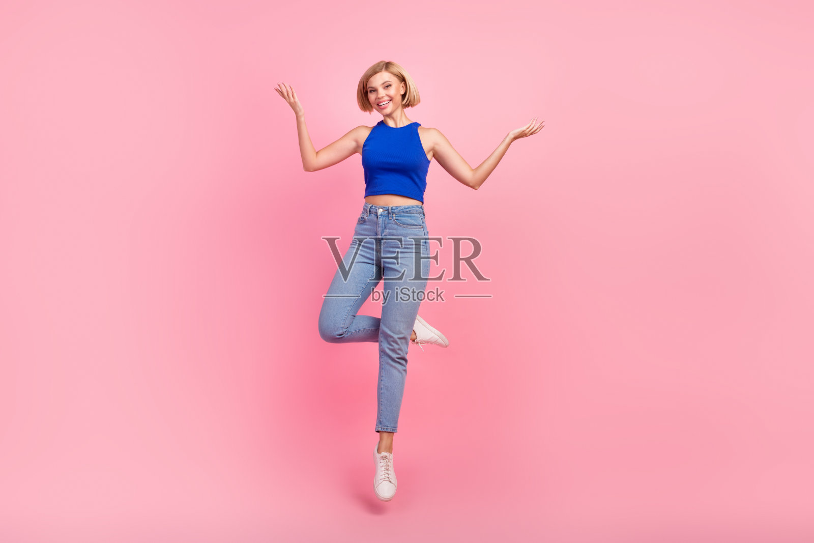 漂亮女孩的全身肖像跳跃手臂显示空的空间穿蓝色上衣孤立在粉红色的背景照片摄影图片