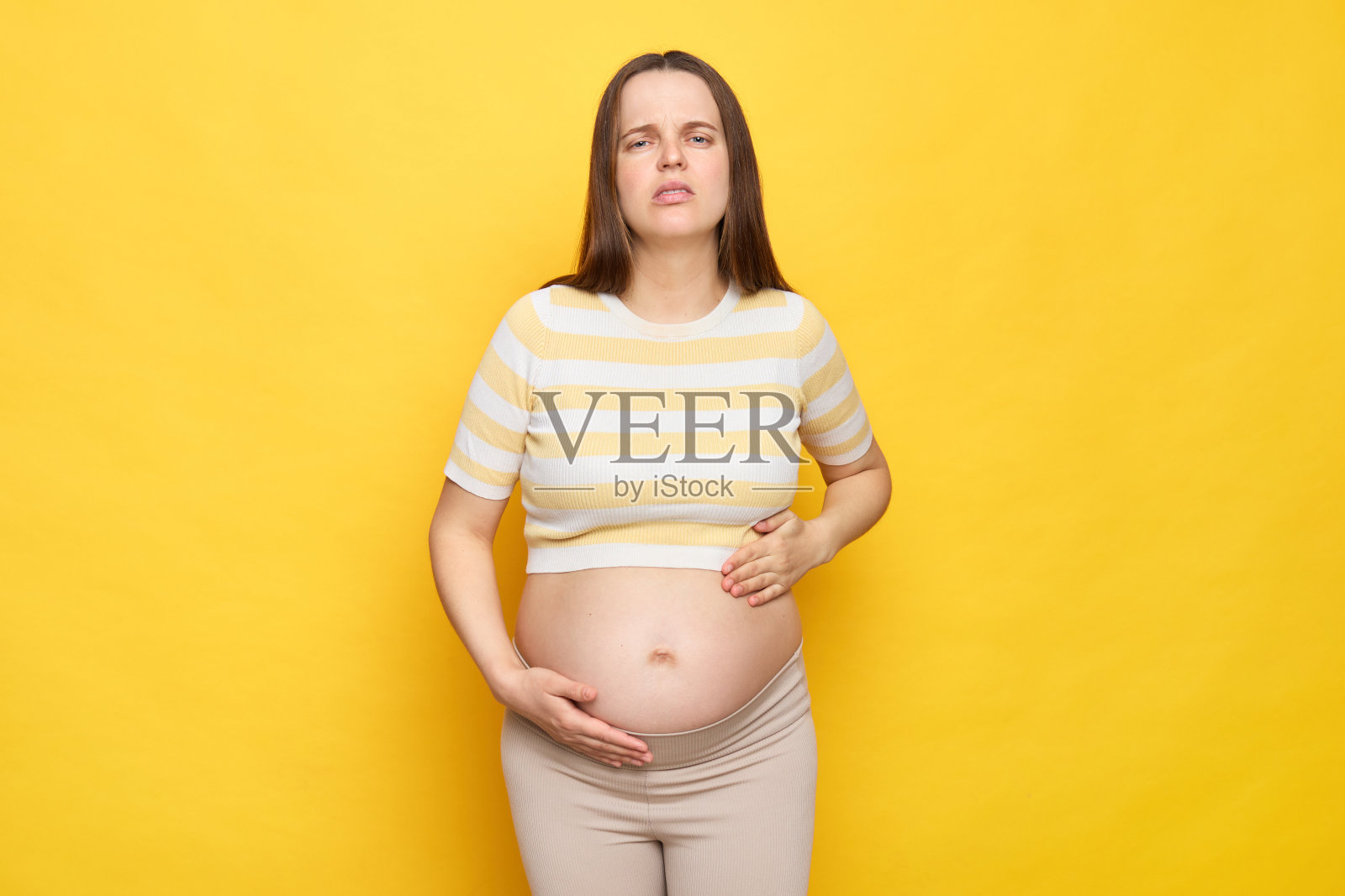 生病的白人孕妇穿着休闲服，在黄色背景上独自摆姿势，感到不适，用针夹住肚子，做着鬼脸摸着肚子照片摄影图片