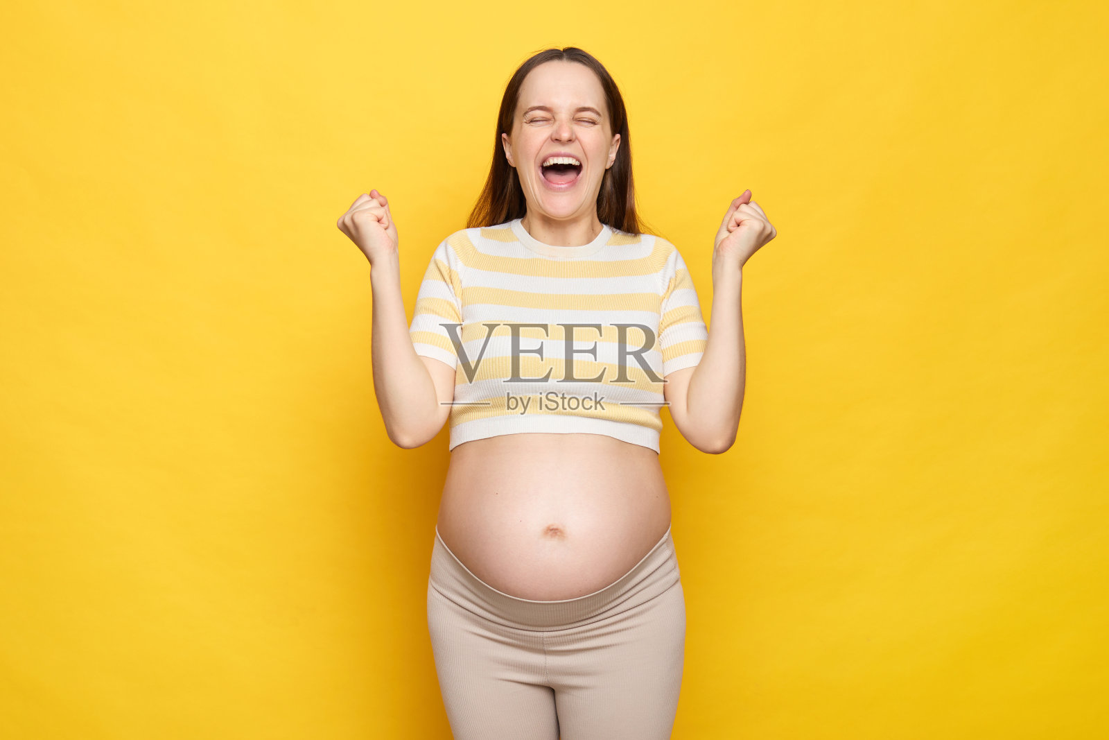 非常高兴，喜出望外的白人孕妇裸露腹部，穿着休闲上衣，孤立在黄色背景下，握紧拳头庆祝她成功的超声波检查照片摄影图片