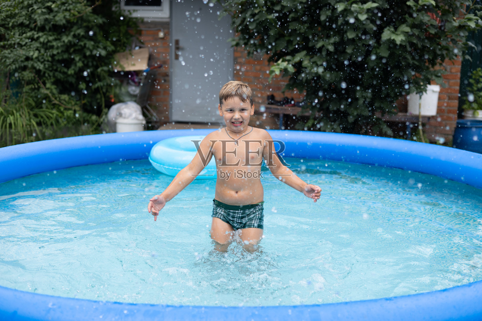 夏日里，一个孩子把泳池里的水泼向镜头。一个漂亮的小男孩在他家后院的游泳池里游泳。夏天的概念，孩子在游泳池游泳。照片摄影图片