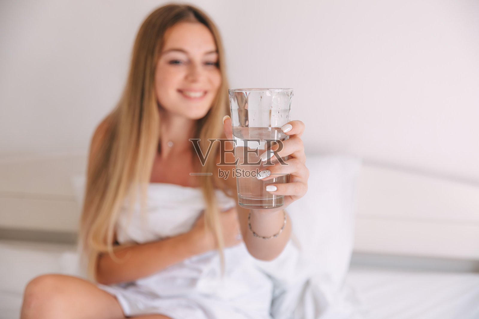快乐的美女早上醒来时在卧室喝水。微笑的女孩在床上拿着透明的玻璃。照片摄影图片