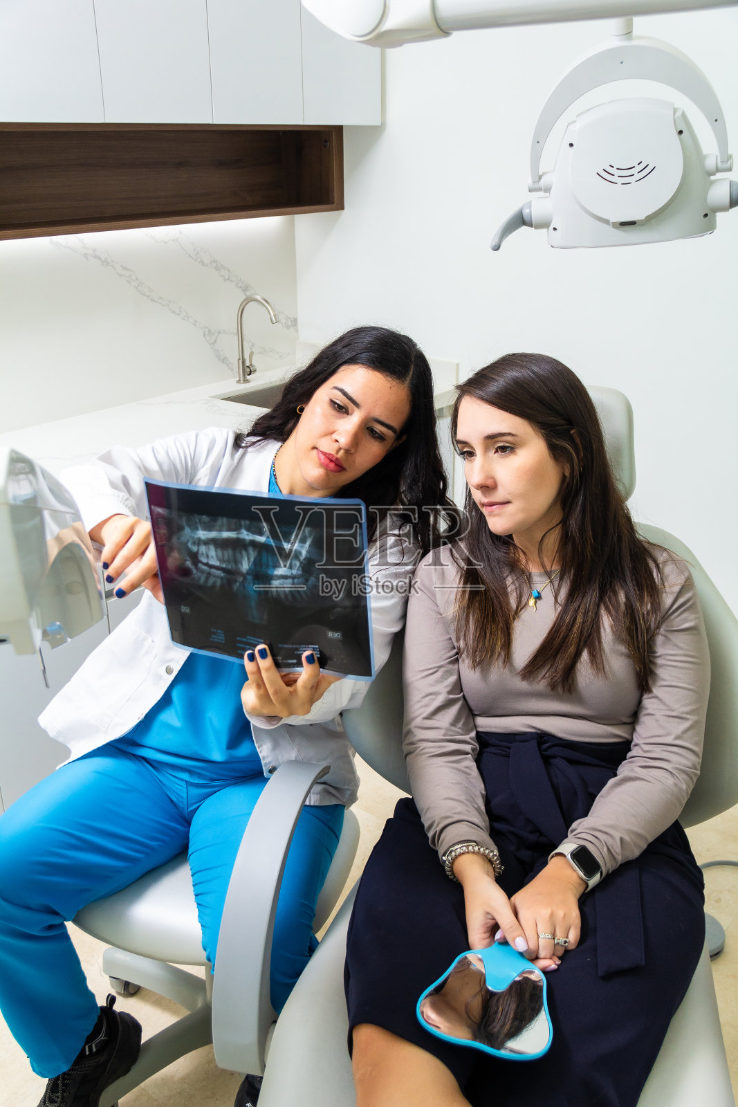 拉丁女牙医向坐在牙科椅上的白人女病人解释牙齿x光图像。牙科保健概念。照片摄影图片