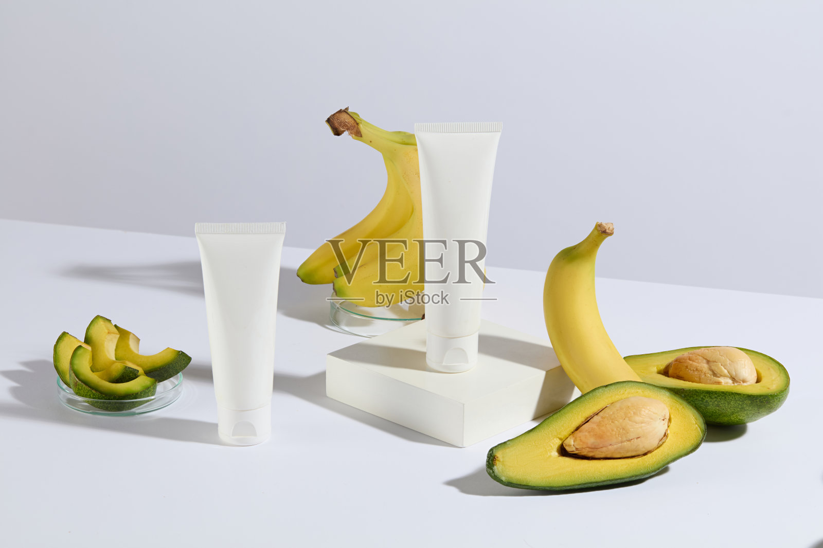 白色色调的牛油果和香蕉组成的照片，两根没有标签的化妆管陈列在桌子上，其中一根放在一个白色的矩形平台上，周围摆放着新鲜的水果照片摄影图片