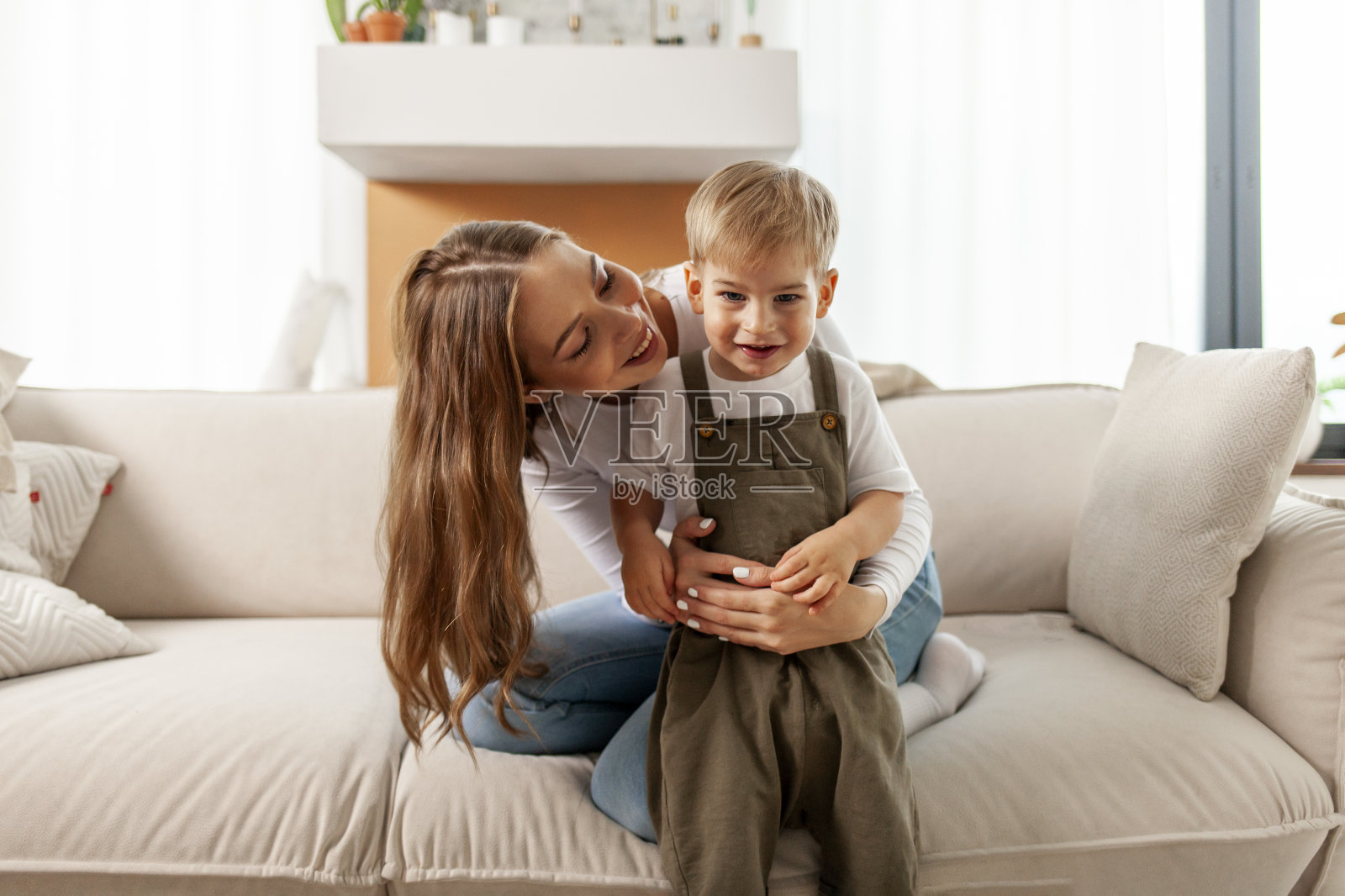 年轻的母亲在沙发上关心和拥抱她的儿子，妇女在家里和她的孩子玩耍，2岁的男孩高兴照片摄影图片