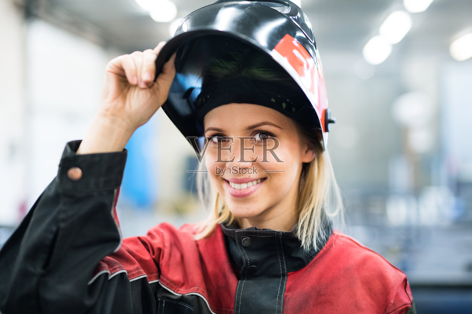 美丽的金发女郎在车间做焊工，操作焊机，穿着防护服，戴着焊接口罩。照片摄影图片