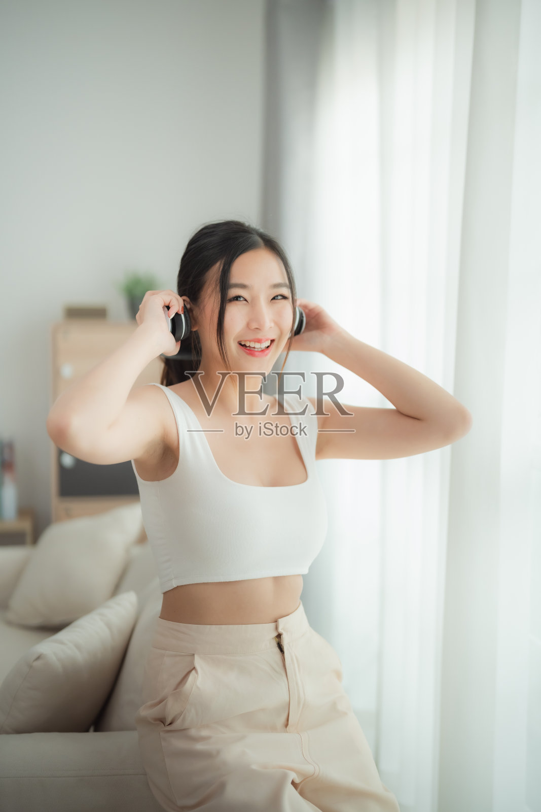 一位快乐的年轻女子，留着长长的黑发，穿着白色露脐上衣和米色裤子，在调整耳机时微笑着。她在明亮、现代的客厅里，洋溢着幸福和活力。照片摄影图片