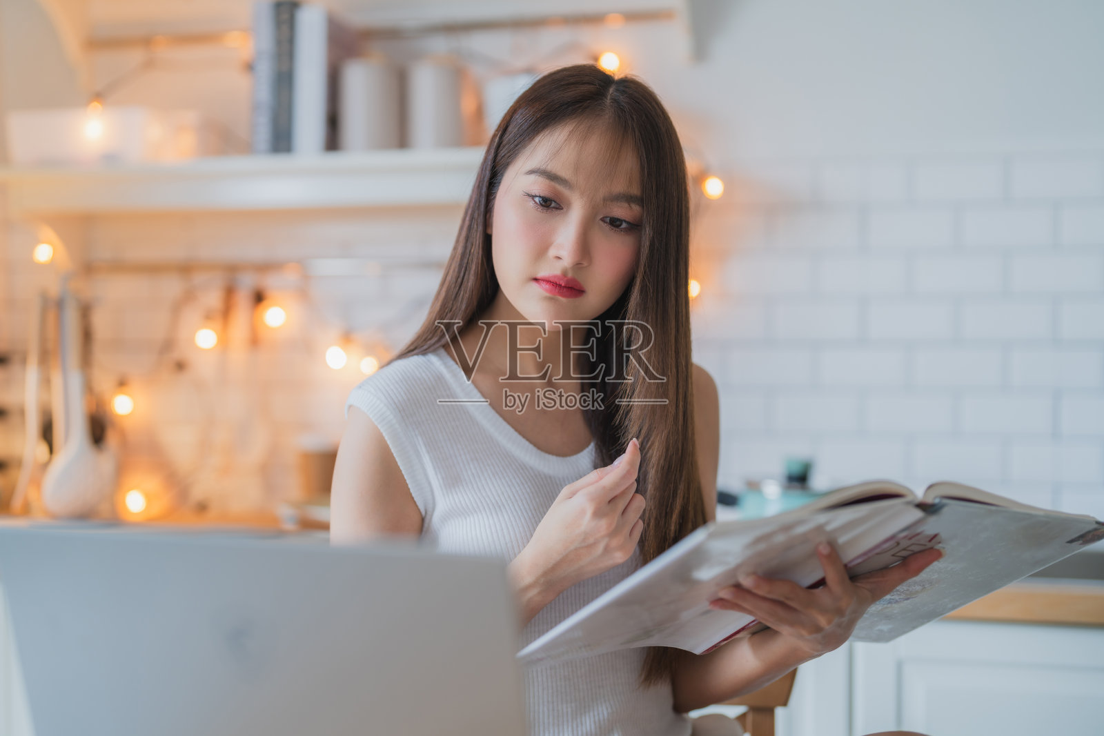 年轻女子坐在桌边看着她的笔记本电脑，喝着咖啡。女性在家里使用笔记本电脑的照片，一位年轻女性在厨房厨房工作时使用笔记本电脑并喝咖啡照片摄影图片