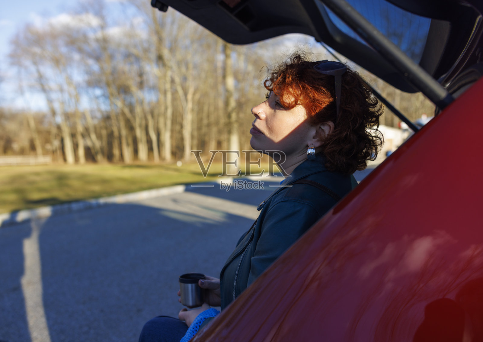春天公路旅行休息。红发女人在停车场汽车后备箱的阴影下享受咖啡休息时间照片摄影图片