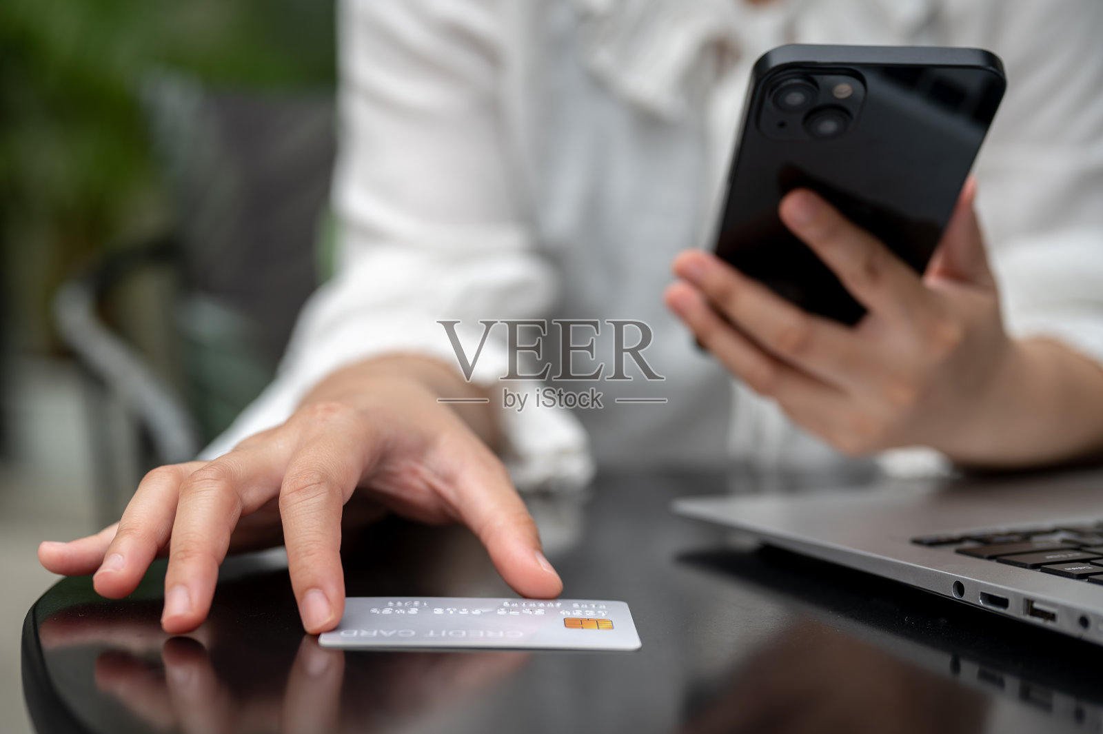 一位女士正在触摸桌子上的信用卡，看着她的信用卡号码，并使用她的手机。照片摄影图片