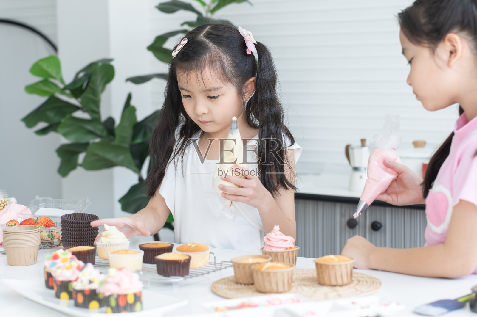 亚洲的两个孩子喜欢在家里的厨房一起做纸杯蛋糕。可爱的小女孩们从装饰袋里挤出奶油来装饰自制的纸杯蛋糕，微笑着参加节日活动照片摄影图片
