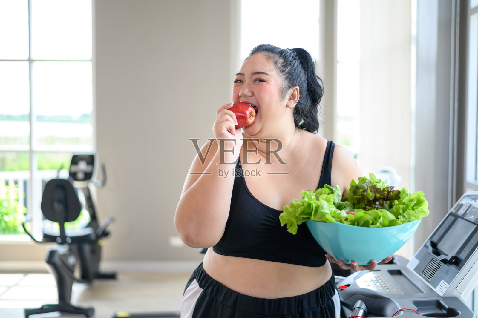 超重的亚洲女性乐于在健身房吃有机苹果。减肥锻炼，健康的生活方式理念。照片摄影图片