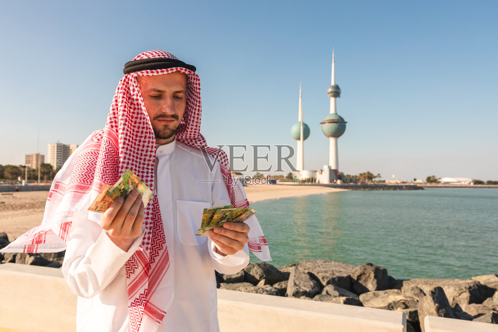 手持钞票的年轻阿拉伯人，背景是科威特塔照片摄影图片