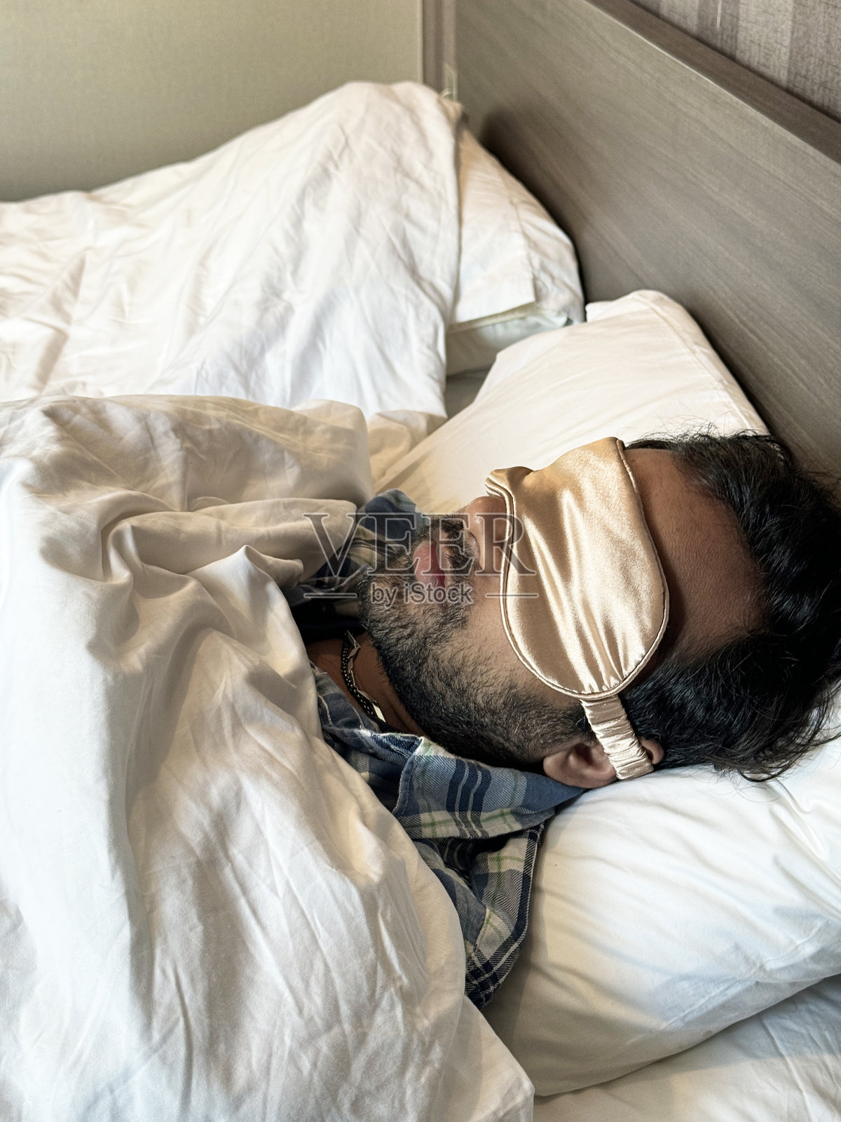 穿着睡衣的印度男子躺在双人床上的枕头上，戴着金色的睡眠眼罩，裹在白色羽绒被下，抬高视野，聚焦于前景照片摄影图片